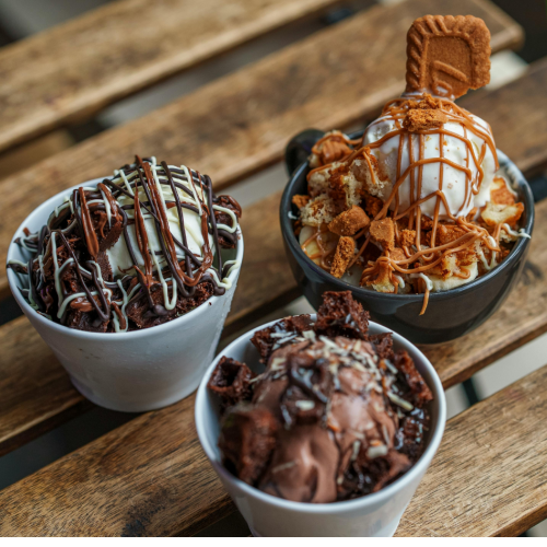 Three bowls of decadent ice cream with various toppings are displayed on a rustic wooden table.