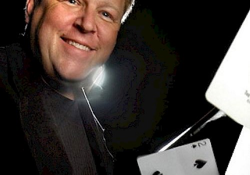 A smiling man wearing a tuxedo has playing cards and a white device in front of him, possibly demonstrating magic or a card trick.