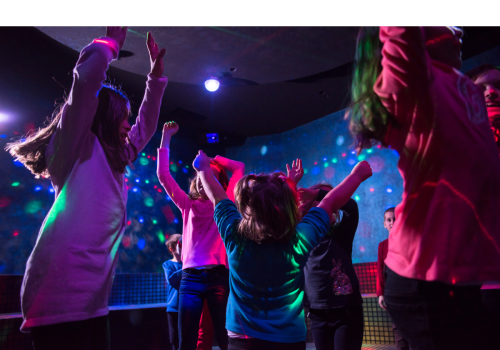 Children are dancing and having fun in a colorful, vibrant indoor setting with lights and a lively atmosphere.