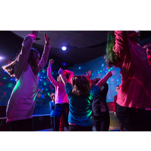 Children are dancing and having fun in a colorful, vibrant indoor setting with lights and a lively atmosphere.