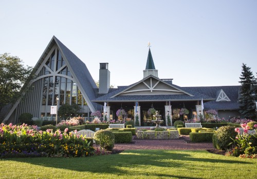 A charming garden with a fountain and benches in front of a whimsical building featuring a steep roof and a tower, surrounded by lush flowers.