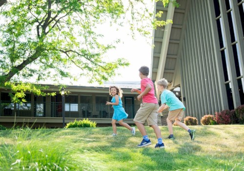 Three kids are playing and running on a grassy yard near a house, enjoying a sunny day in a lush, green outdoor setting.