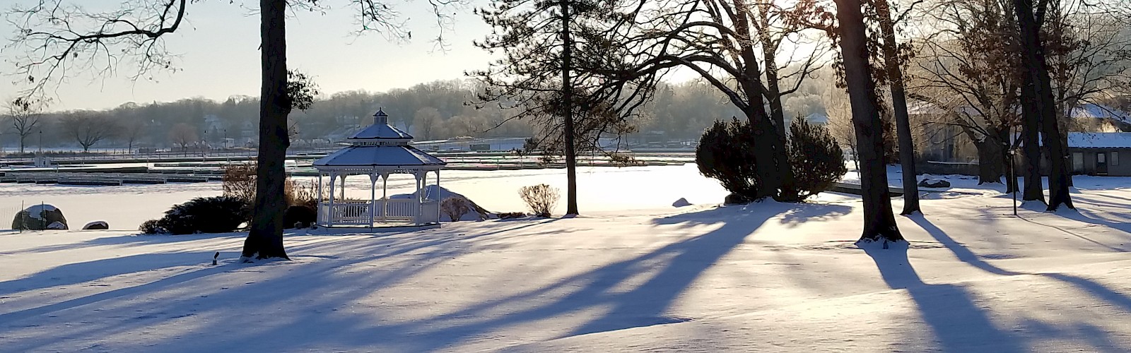 A snowy park scene at sunrise with trees, a gazebo, and long shadows on the snow.