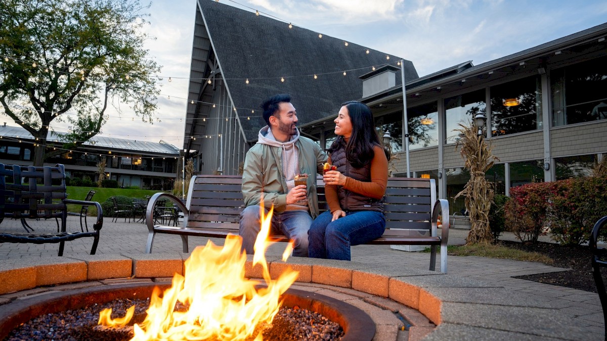 A couple sits on a bench near a fire pit, enjoying drinks and chatting outside at dusk.
