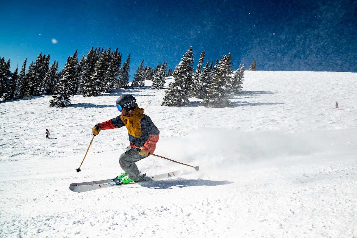 A skier in colorful gear glides downhill on a snowy mountain with pine trees and other skiers in the background under a bright blue sky.