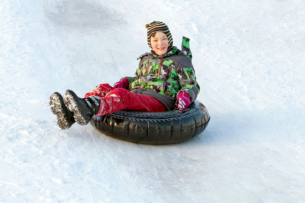 A child wearing colorful winter clothes is smiling while riding a snow tube down a snowy slope.