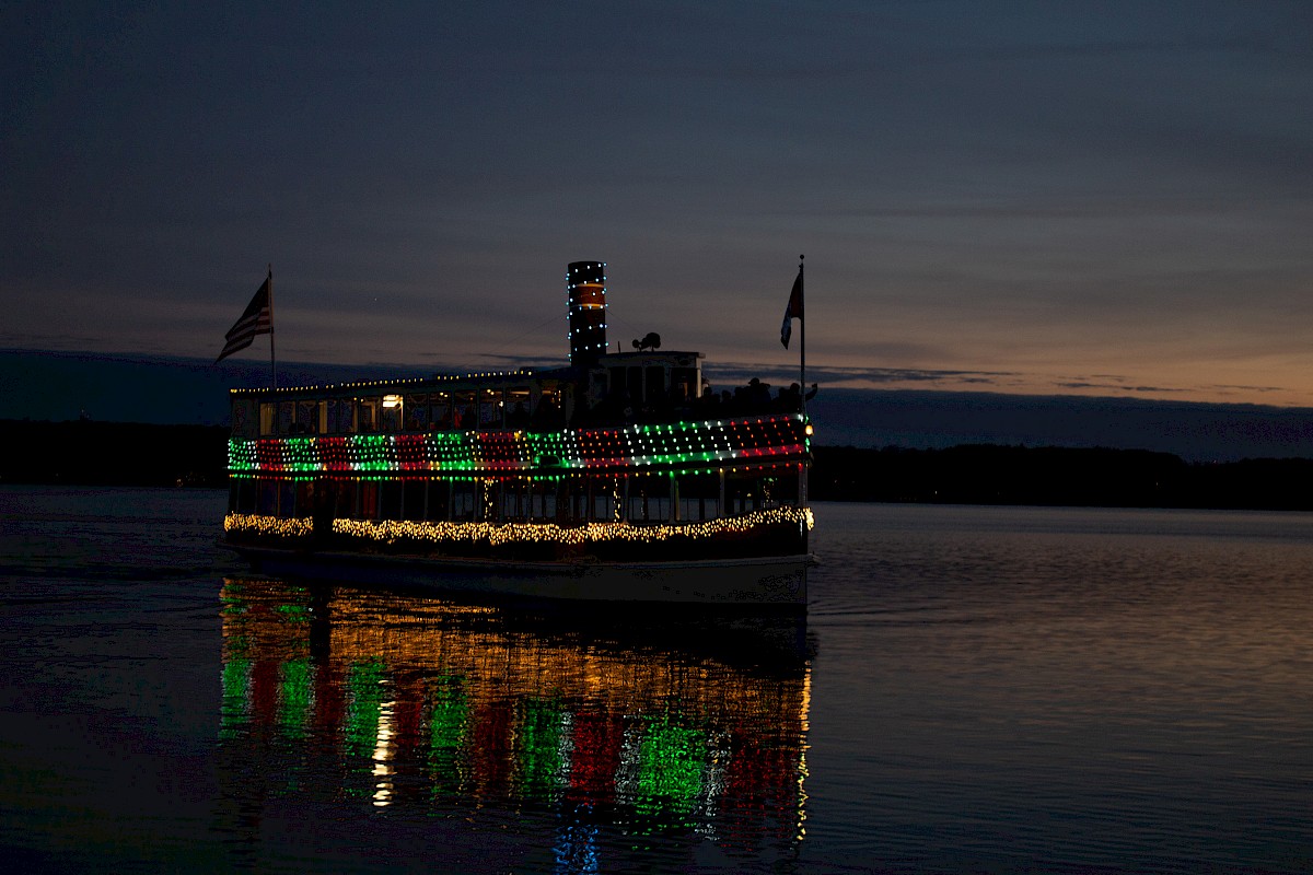 A boat decorated with Christmas lights is sailing on a calm river at sunset with colorful reflections on the water.