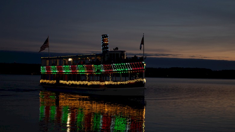 A boat decorated with Christmas lights is sailing on a calm river at sunset with colorful reflections on the water.