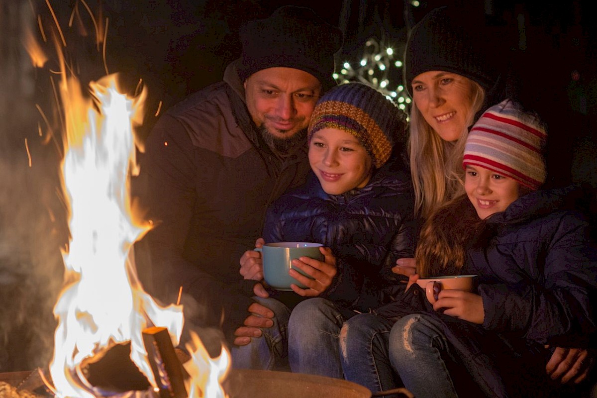 A family of four, bundled up in warm clothes, enjoys sitting around a campfire at night, sharing smiles and hot drinks.