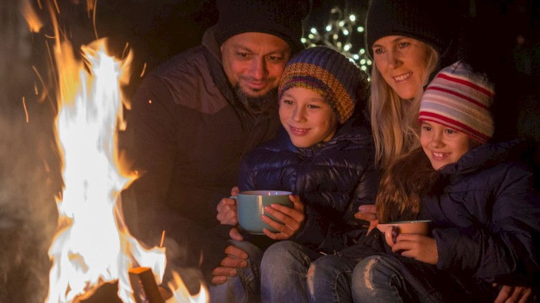 A family of four, bundled up in warm clothes, enjoys sitting around a campfire at night, sharing smiles and hot drinks.