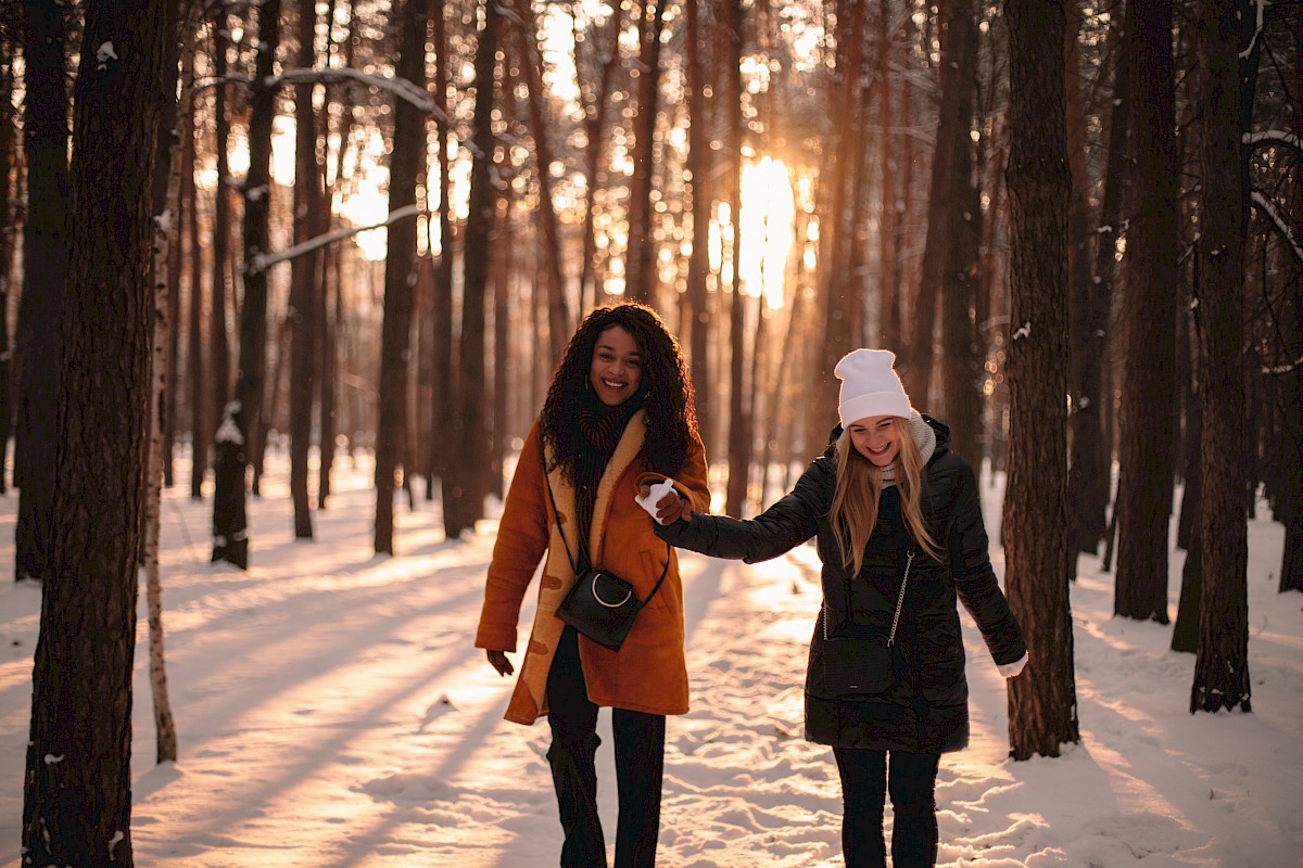 Two women are smiling and holding hands while walking in a snowy forest during sunset.