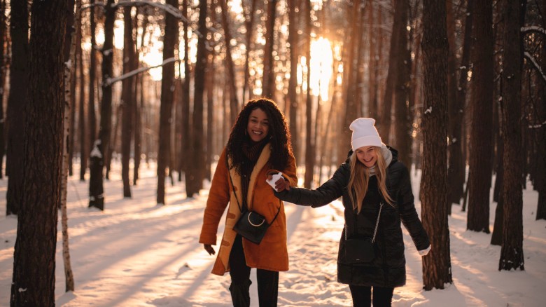 Two women are smiling and holding hands while walking in a snowy forest during sunset.