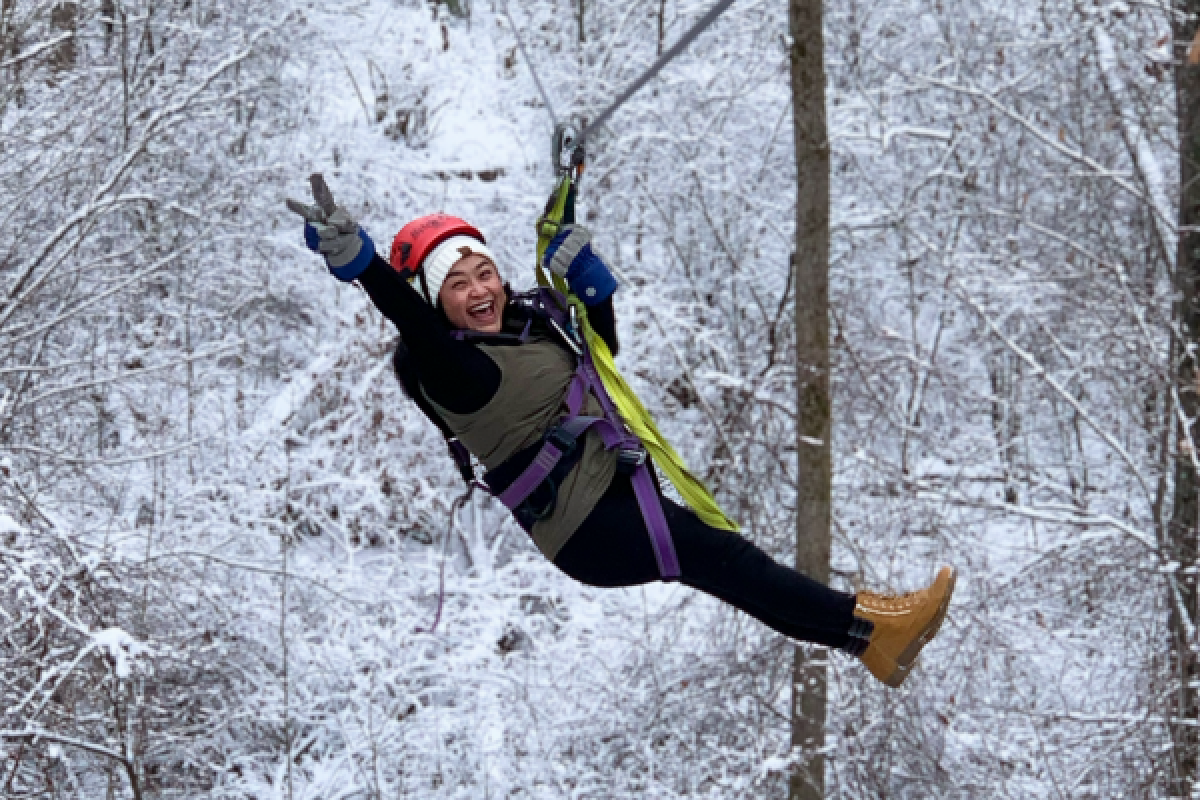 A woman wearing a helmet and harness is zip-lining through a snowy forest with a joyful expression.