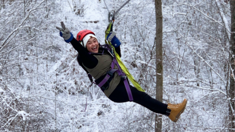 A woman wearing a helmet and harness is zip-lining through a snowy forest with a joyful expression.