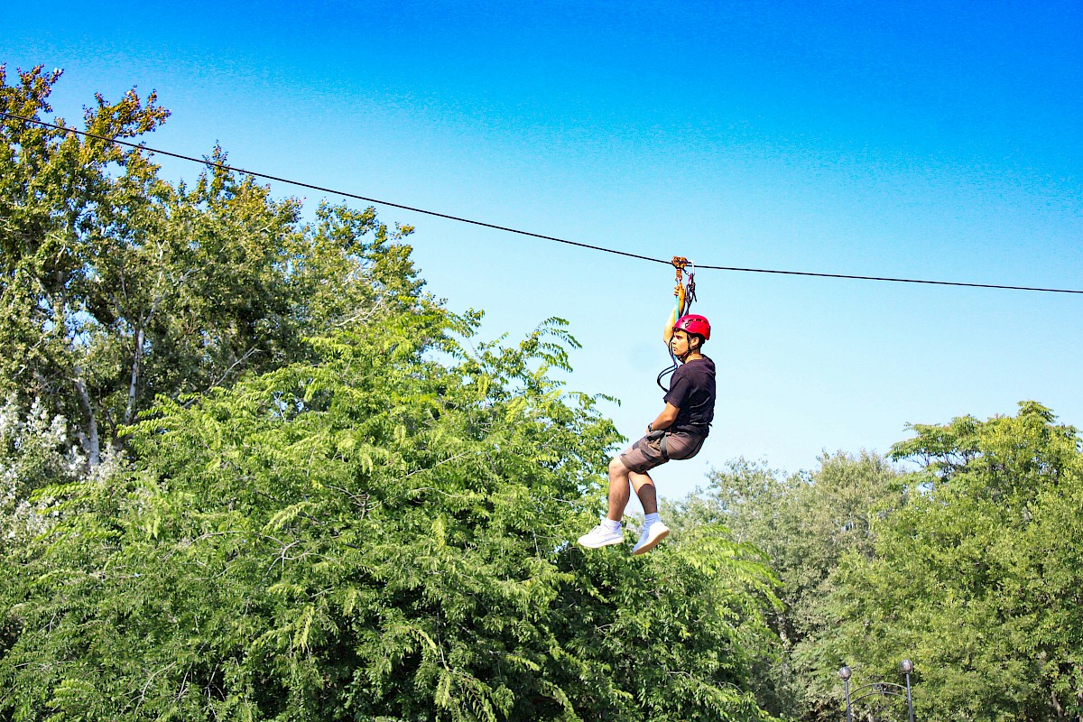 A person wearing a red helmet is riding a zip line above green trees on a sunny day.