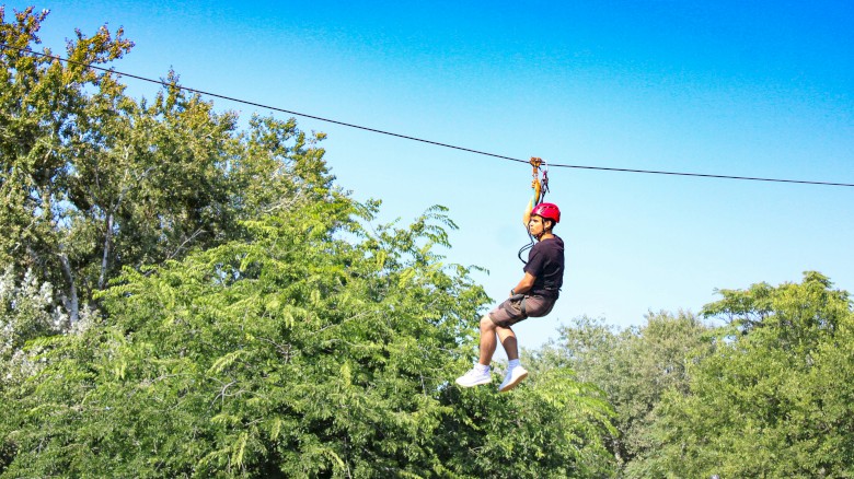 A person wearing a red helmet is riding a zip line above green trees on a sunny day.
