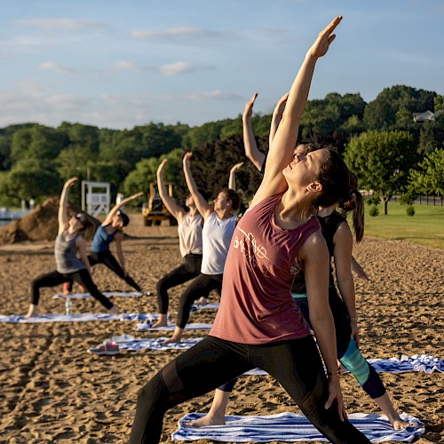 Women practicing outdoor yoga on beach towels during sunset, with trees and hills in the background.