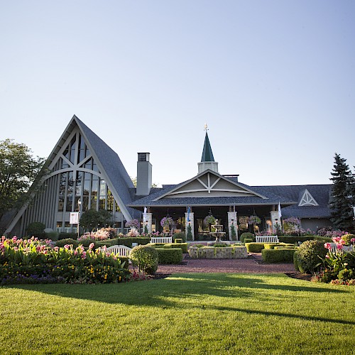 A charming garden with a fountain and benches in front of a whimsical building featuring a steep roof and a tower, surrounded by lush flowers.