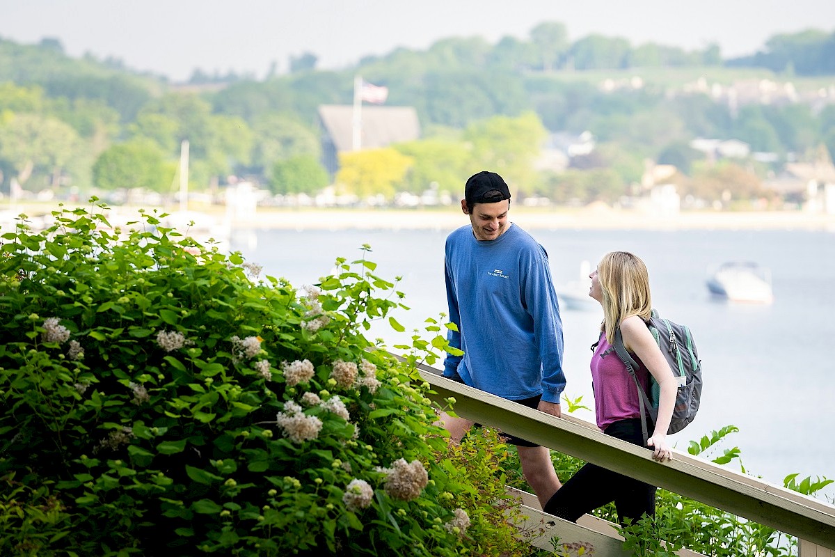 A young man and woman are talking by a waterfront, surrounded by greenery, with boats and distant hillside homes in the background.