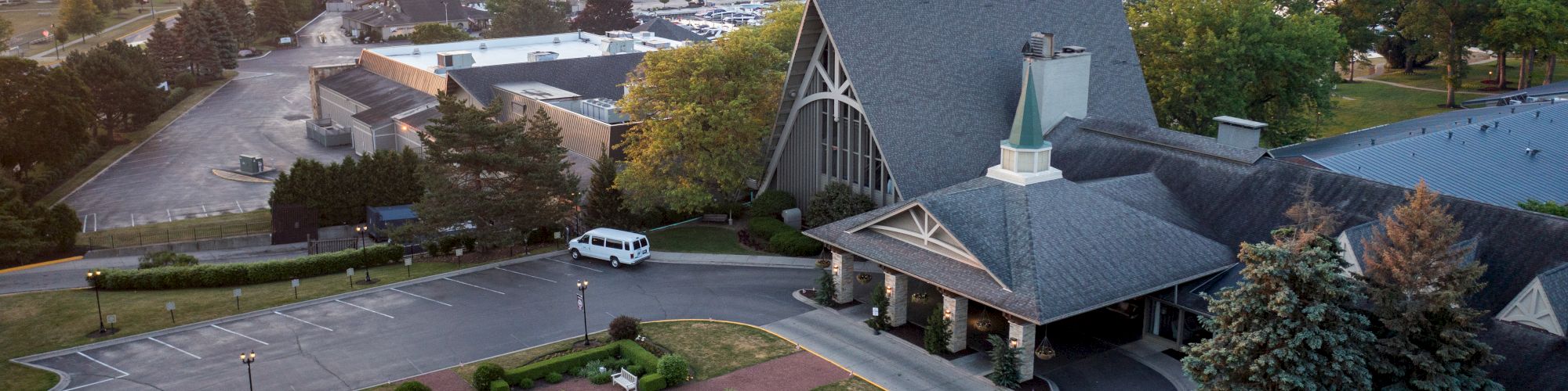 An aerial view shows a building with a manicured garden, nearby parking lot, and distant water body with surrounding greenery at sunset.