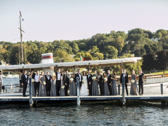 A group of people dressed formally standing on a dock by the water with a boat to their left and a green, wooded area in the background.