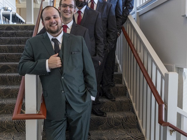 Five men in suits and ties stand on a staircase, smiling at the camera. The setting appears to be formal, possibly for an event or gathering.