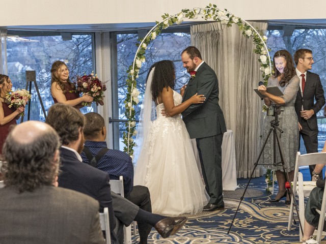 A couple is exchanging vows under an arch with a floral arrangement. The bride wears a white gown, and the groom is in a dark suit.