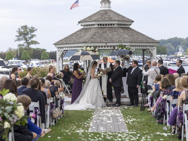 A wedding ceremony is taking place outdoors under a gazebo with guests seated around, facing a bride and groom with umbrellas.