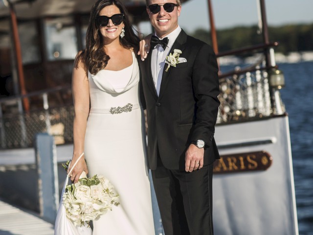 A smiling bride and groom in formal attire stand on a dock by a boat, holding flowers under a clear sky. The boat’s name is partially visible in the background.