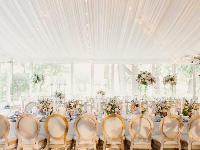A beautifully decorated event space with elegant chairs, floral centerpieces, and string lights hanging from a draped ceiling.