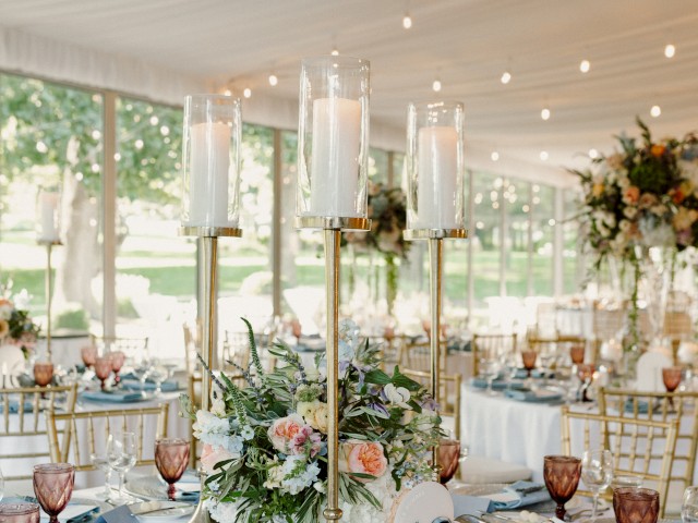 Elegant event setup with dressed tables featuring tall candle holders, floral arrangements, and ornate tableware under a draped ceiling.