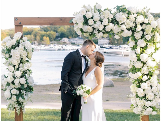 A couple is kissing under a floral arch at an outdoor wedding ceremony by the water, with a picturesque background of boats and buildings.