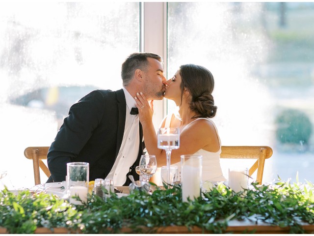 A couple dressed in formal attire is kissing at a beautifully decorated table with green foliage and candles, inside a sunlit room.