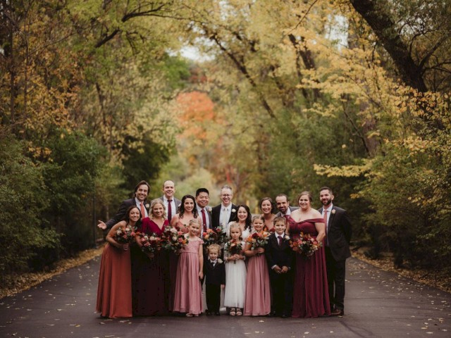 A wedding party is posing on a wooded, scenic road with people dressed in formal attire, featuring bridesmaids and groomsmen.