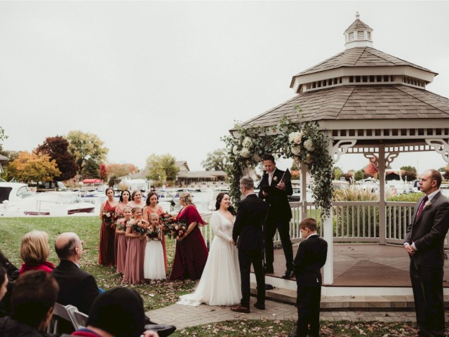A wedding ceremony at a gazebo with the bride, groom, bridal party, and guests under cloudy skies near a lake with autumn trees.
