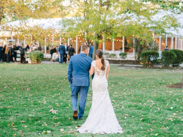 A couple, with the woman in a wedding dress, walks towards a gathering under a tent in a scenic outdoor setting.