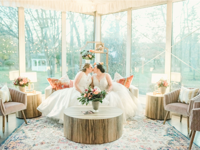 Two brides in wedding dresses share a kiss in a beautifully decorated room with floral arrangements, cozy chairs, and a round wooden table.