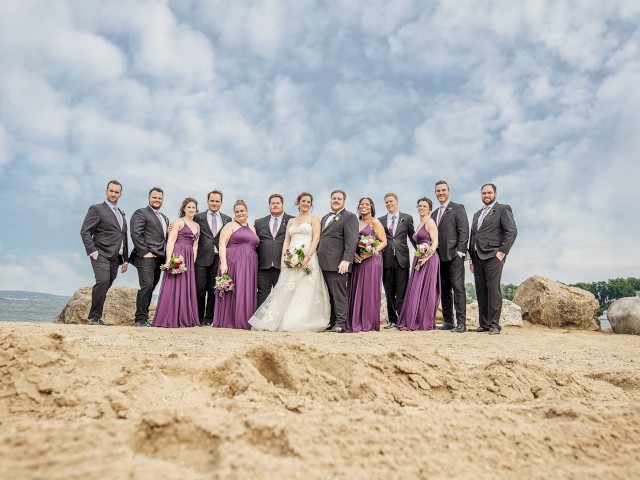 A wedding party is posing on a beach with bridesmaids in purple dresses and groomsmen in dark suits, with the ocean and sky in the background.