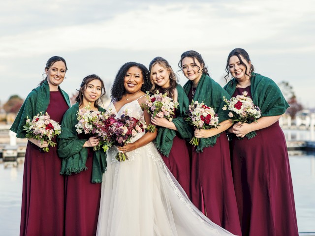 A bride and five bridesmaids in matching dresses and green shawls are posing for a photo by a waterfront. They are all holding bouquets.