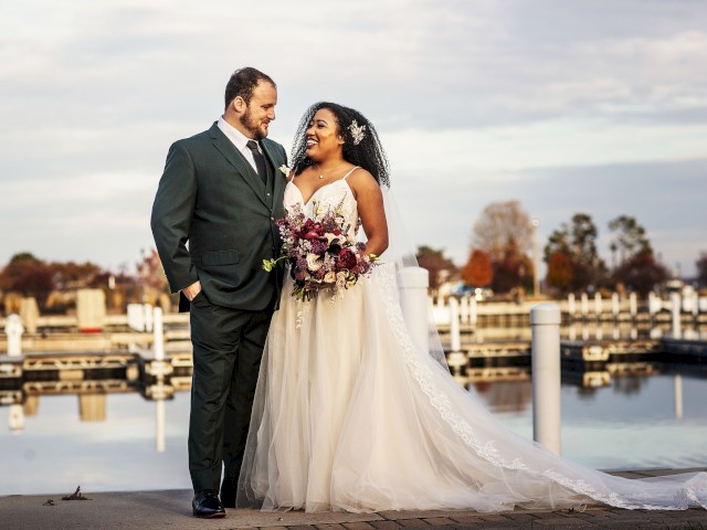 A couple stands by a waterfront wearing wedding attire. The bride holds a bouquet, and they smile at each other with boats and a marina in the background.
