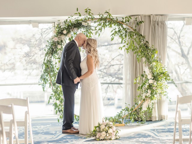 A couple is standing under a circular floral arch, holding hands and leaning in for a kiss at what appears to be a wedding ceremony.