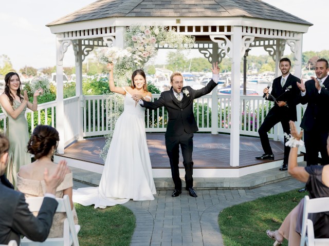 Newlyweds stand under a gazebo with raised hands, surrounded by applauding guests and bridesmaids in a garden setting, concluding their ceremony.