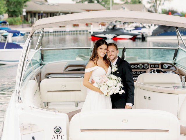 A couple in wedding attire is standing on a boat, posing for a photo with a marina in the background. The boat's name is 