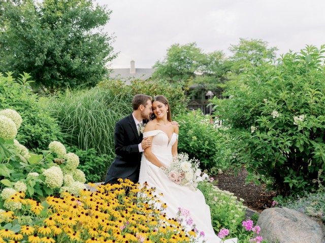 A bride and groom are sitting in a lush garden surrounded by vibrant flowers and greenery, sharing an intimate moment.