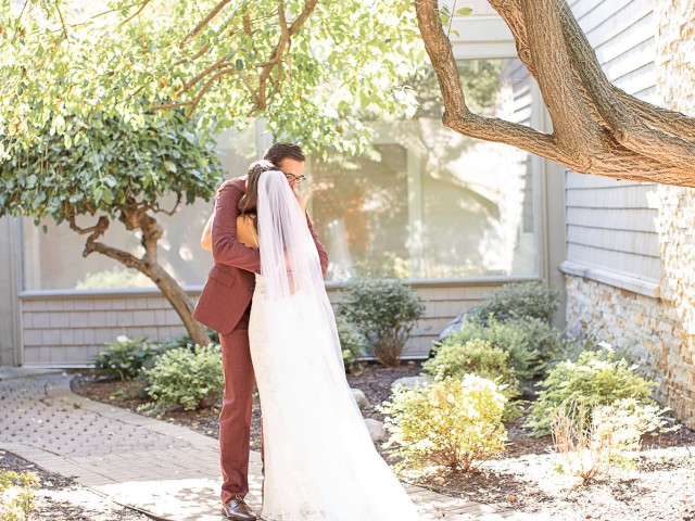 A bride and groom stand under a tree, embracing in a garden setting with surrounding greenery and sunlight filtering through.