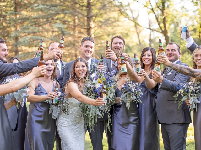 A group of elegantly dressed people, likely a wedding party, are toasting with bottled beverages and smiling outdoors in a wooded area.
