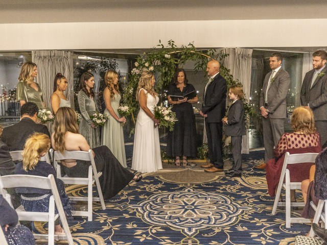 A wedding ceremony with a couple exchanging vows, an officiant, bridesmaids, groomsmen, and guests seated, all set in an indoor venue.