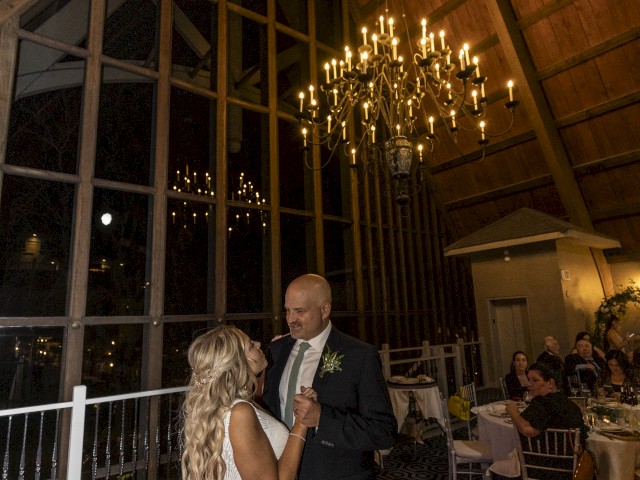 A couple is dancing in a large hall with a chandelier and guests seated at tables, reflecting a wedding celebration under dim lighting conditions.