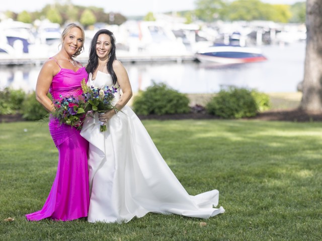 Two women pose together, one in a white wedding dress and the other in a bright pink gown, holding bouquets, near a marina with boats in the background.