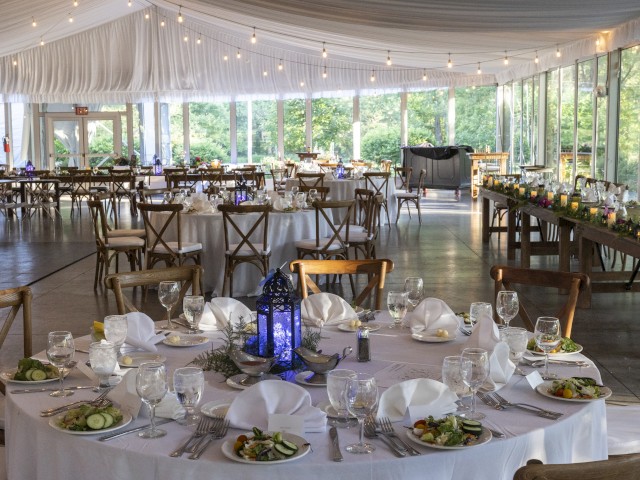An elegant, decorated event space with round tables set for dining, adorned with napkins, glasses, and salads, under string lighting.
